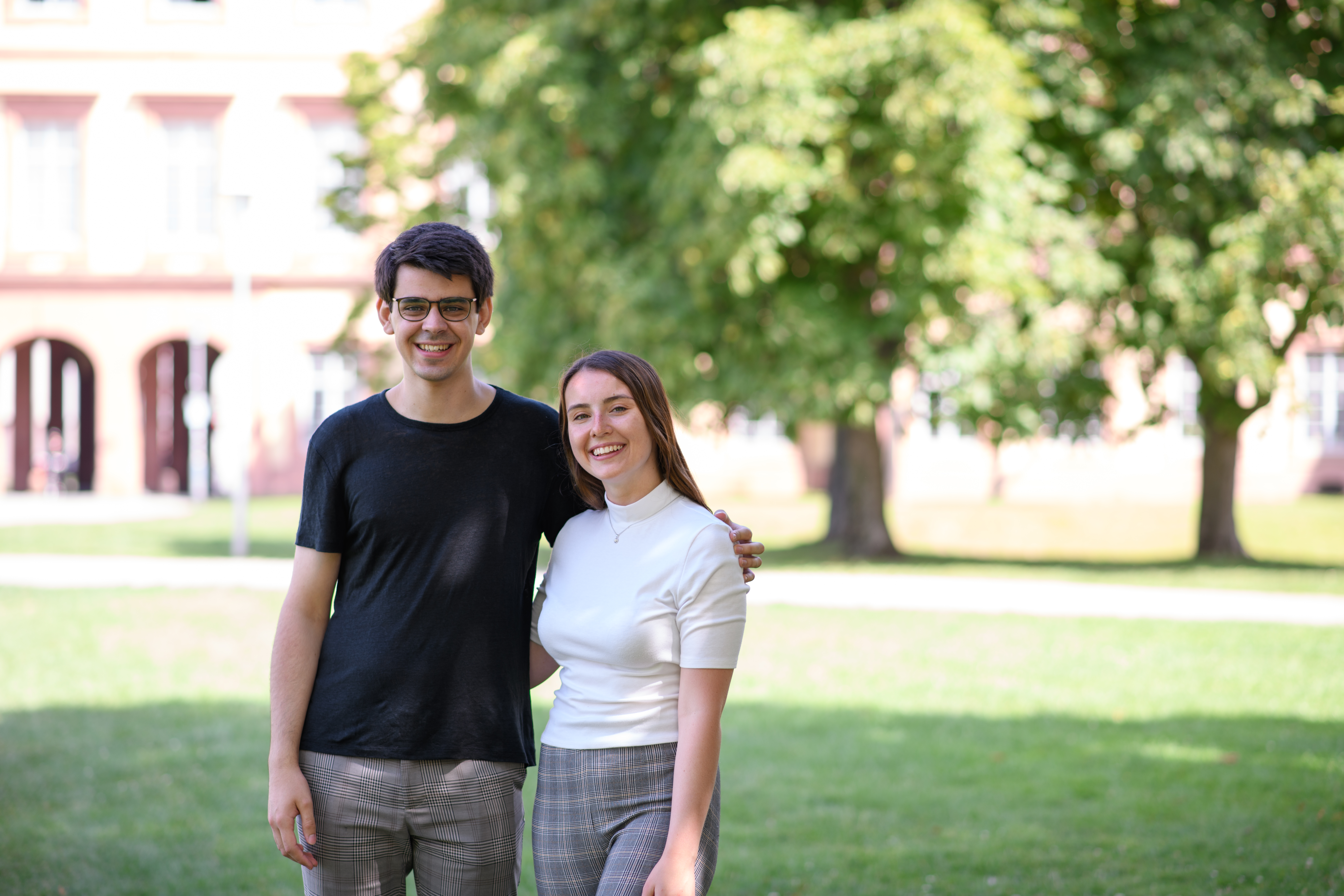 Zwei lächelnde Studierende stehen auf der grünen Mensawiese vor dem Schloss