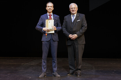 Auf einer Bühne vor schwarzem Hintergrund stehen auf der linken Seite Max Spanier mit dem Pfalzpreis und rechts der Bezirkstagsvorsitzende Hans-Ulrich Ihlenfeld. 
