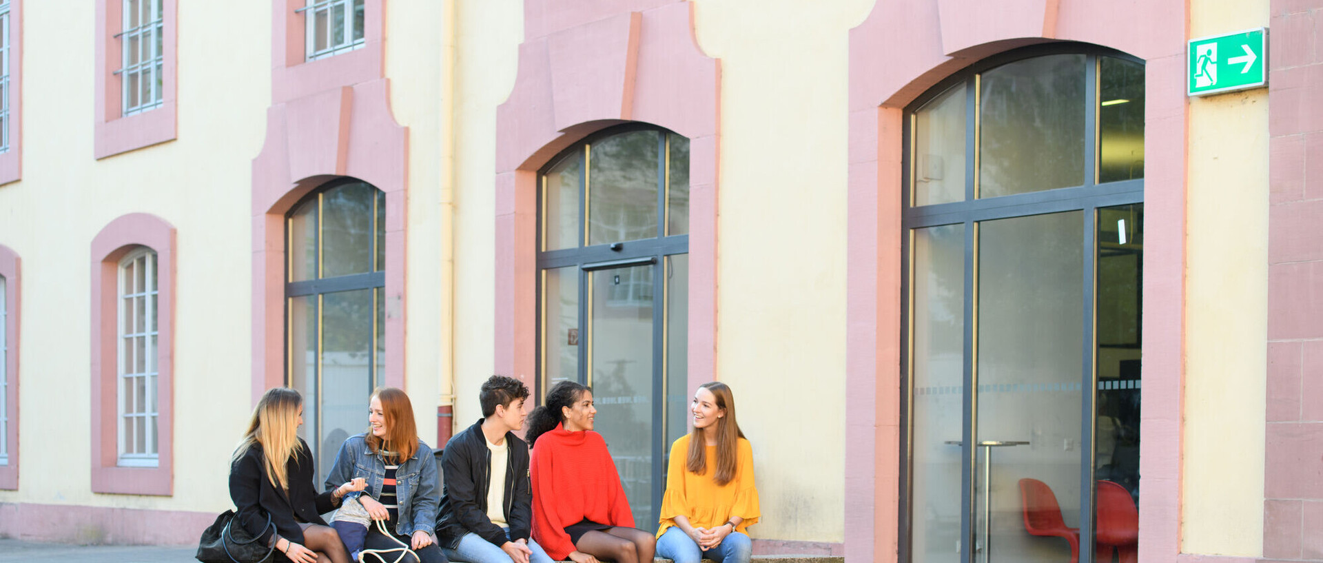 Fünf Studierende sitzen im Schneckenhof auf einer Mauer vor der Bibliothek. 