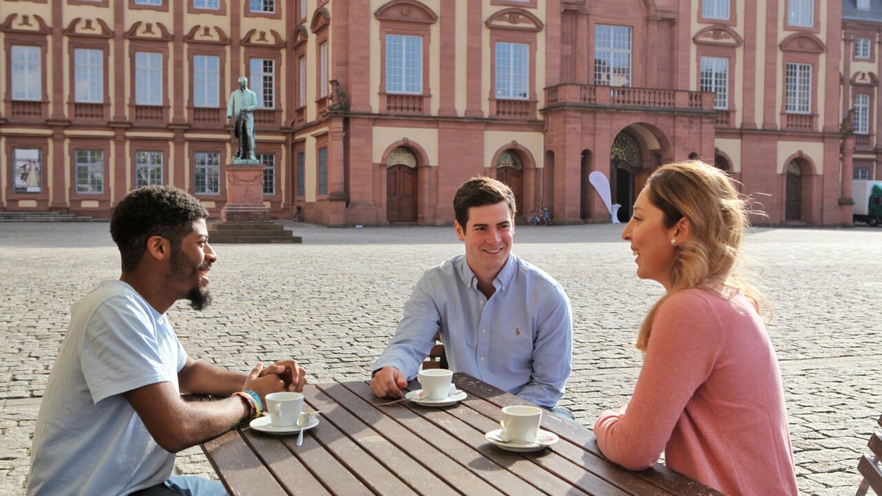 Zwei junge Männer und eine junge Frau sitzen an einem Holztisch auf dem Ehrenhof. Vor ihnen stehen drei weiße Tassen.
