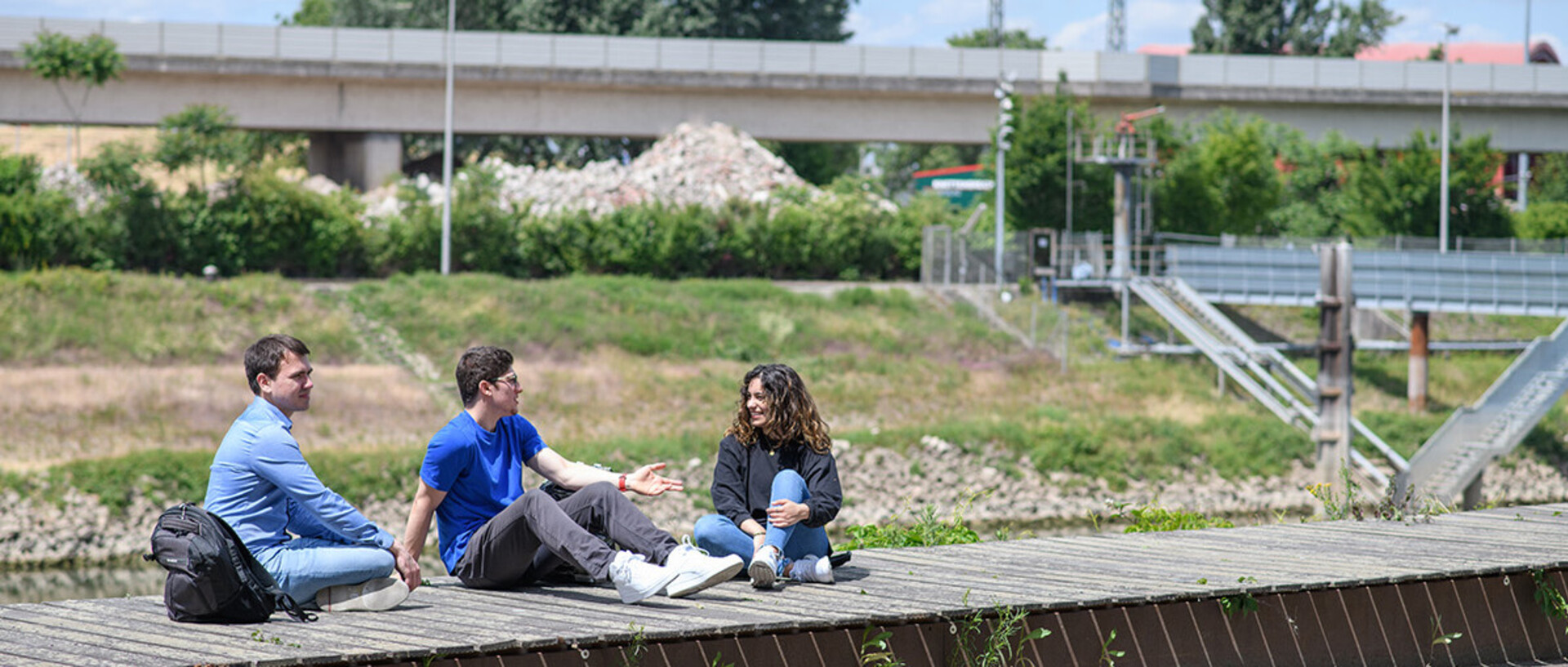 Drei Studierende sitzen in der Sonne auf einem Holzsteg. Im Hintergrund eine Brücke.