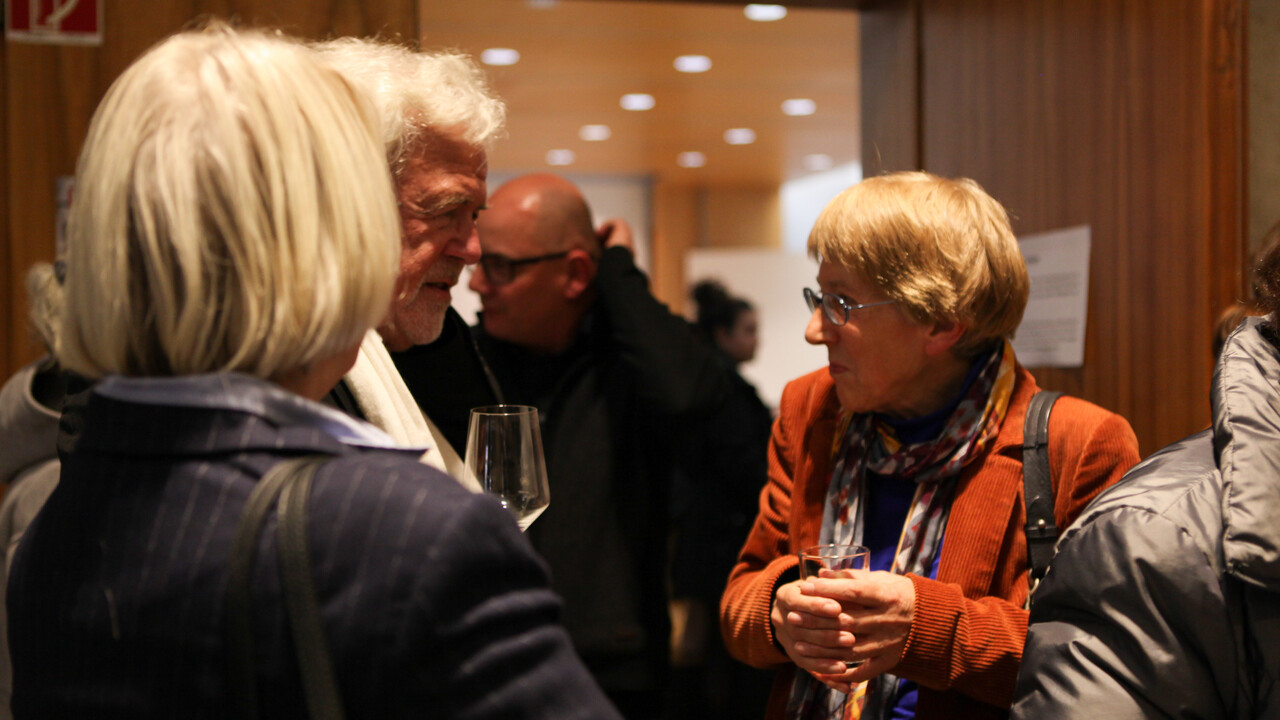 Einige Personen stehen winterlich mit Jacken und Anzügen bekleidet mit Gläsern in den Händen in einem holzvertäfelten Ambiente. Identifizierbar ist Prof. Dr. Ursula Wolf im Gespräch.