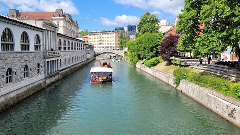 Malerischer Blick auf den Fluss mit einem Boot, das unter einer Steinbrücke hindurchfährt, flankiert von einem historischen Gebäude und üppig grünen Bäumen unter strahlend blauem Himmel.