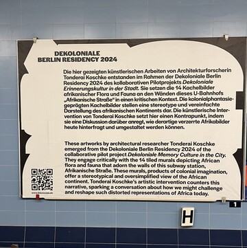 Eine Infotafel in einer Berliner U-Bahnstation. Darauf steht: "DEKOLONIALE BERLIN RESIDENCY 2024. Die hier gezeigten künstlerischen Arbeiten von Architekturforscherin Tonderai Koschke entstanden im Rahmen der Dekoloniale Berlin Residency 2024 des kollaborativen Pilotprojekts Dekoloniale Erinnerungskultur in der Stadt. Sie setzen die 14 Kachelbilder afrikanischer Flora und Fauna an den Wänden dieses U-Bahnhofs „Afrikanische Straße" in einen kritischen Kontext. Die kolonialphantasie-geprägten Kachelbilder stellen eine stereotype und vereinfachte Darstellung des afrikanischen Kontinents dar. Die künstlerische Intervention von Tonderai Koschke setzt hier einen Kontrapunkt, indem sie eine Diskussion daüber anregt, wie derartige verzerrte Afrikabilder heute hinterfragt und umgestaltet werden können."