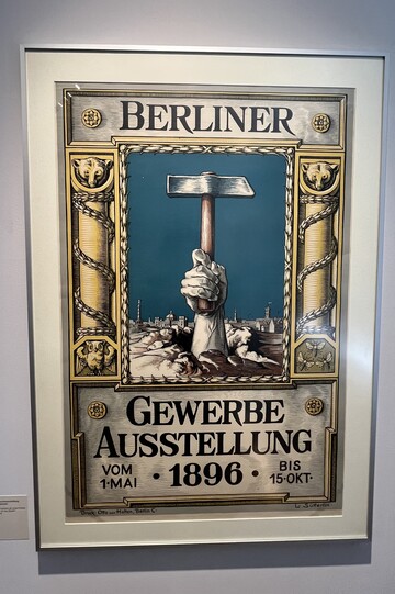 Ein Plakat für die erste Berliner Gewerbeausstellung 1896. Die Gestaltung des Plakats suggeriert, dass es sich um eine Metalltafel oder Ähnliches handelt. Abgebildet ist ist in der Mitte eine silberne Hand, die einen Hammer nach oben streckt und aus der Erde hervorbricht, im Hintergrund die Silhouette Berlins. Links und rechts daneben befinden sich goldene Säulen. Über der Abbildung steht "Berlin", darunter "Gewerbeausstellung 1896. Vom 1. Mai bis 15. Okt."