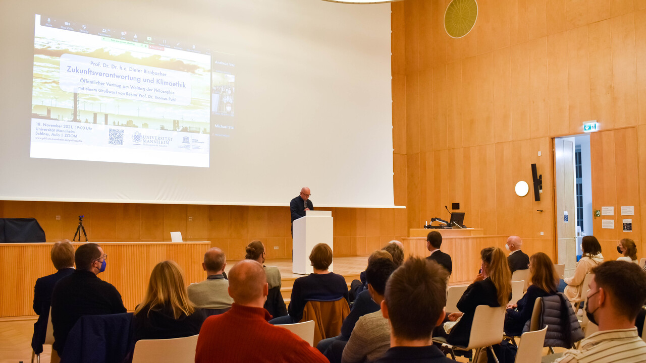 Blick aus dem Publikum auf Prof. Dr. Wolfgang Freitag bei der Begrüßung der Gäste. Man sieht die Begrüßungsfolie der Powerpointpräsentation auf der großen weißen Projektionsfläche in der holzgetäfelten Aula des Schlosses. Prof. Freitag steht in schwarz gekleidet hinter einem Pult auf der leicht erhöhten, teilweise abgetrennten und auf einem Tisch mit einem aufgeklappten Laptop und anderen technischen Geräten versehenen Bühne.