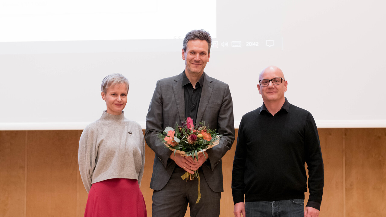 Gruppenfoto: Zentral Redner Prof. Dr. Tobias Rosefeldt mit schwarzem Hemd sowie Jackett und Hose in grau, einen Blumenstrauß in den Händen vor sich haltend. Links davon Dekanin Prof. Dr. Cornelia Ruhe mit hellem Pullover und rotem Rock bei auf den Rücken verschränkten Armen. Rechts der Geschäftsführende Direktor des Philosophischen Seminars Prof. Dr. Wolfgang Freitag in schwarzem Pullover und Jeans mit Brille und herabhängenden Armen. Den Hintergrund des Bildes bilden Holz-Wandvertäfelung der Uni-Aula und helle Projektionsfläche mit projiziertem angeschnittenen weißen Rechteck.