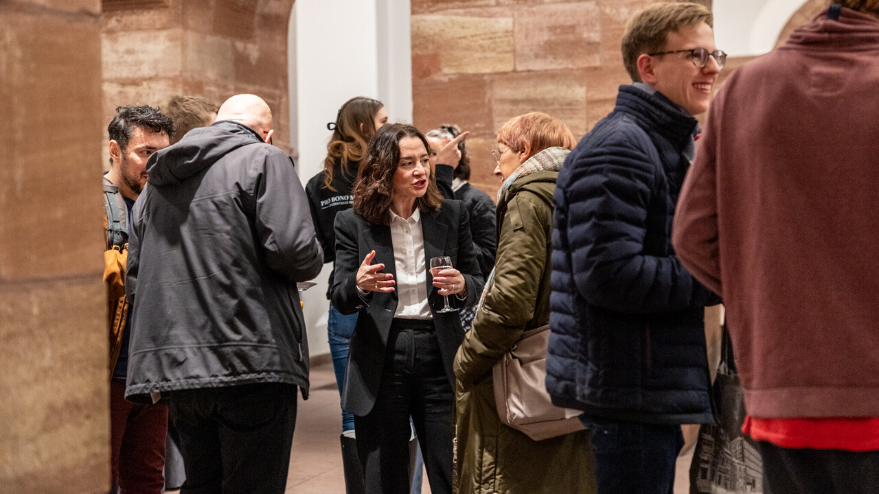 Impressionen vom Empfang in den „Katakomben“ Empfang in den Katakomben der Universität Mannheim am Tag der Philosophie am 20.11.2025: Etwa 1 Dutzend Personen im Gespräch zwischen der Säulen mit Rundbögen aus rotem Sandstein und weißen Wänden im Hintergrund. Zentral im Bild, gekleidet im dunklen Anzug und hellem Hemd mit einem Glas in der Hand Prof. Elif Özmen im Gespräch mit Prof. Ursula Wolf, die einen karierten hellen Schal und eine grüne Jacke mit umgehängter beiger Tasche trägt.
