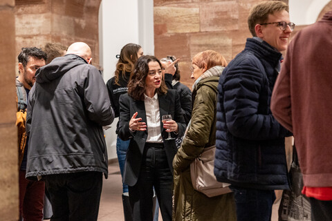 Empfang in den Katakomben der Universität Mannheim am Tag der Philosophie am 20.11.2025: Etwa 1 Dutzend Personen im Gespräch zwischen der Säulen mit Rundbögen aus rotem Sandstein und weißen Wänden im Hintergrund. Zentral im Bild, gekleidet im dunklen Anzug und hellem Hemd mit einem Glas in der Hand Prof. Elif Özmen im Gespräch mit Prof. Ursula Wolf, die einen karierten hellen Schal und eine grüne Jacke mit umgehängter beiger Tasche trägt.