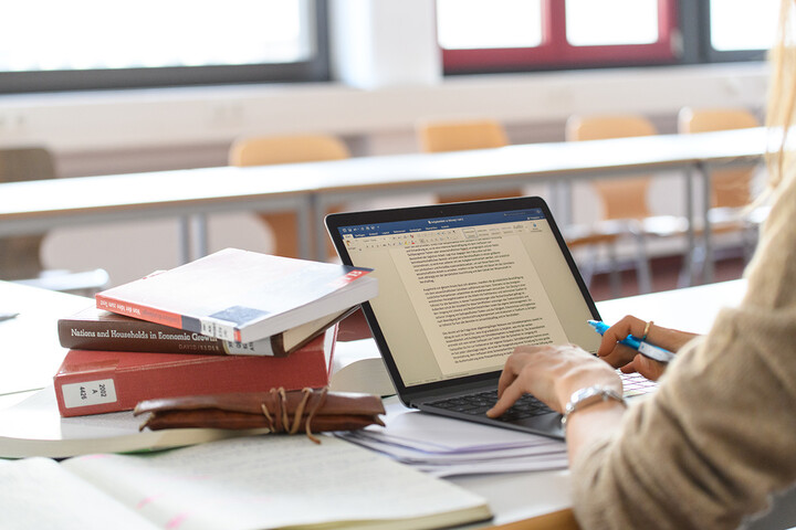 Eine Frau schreibt auf ihrem Laptop in einem Word-Dokument, daneben liegen einige Bücher.