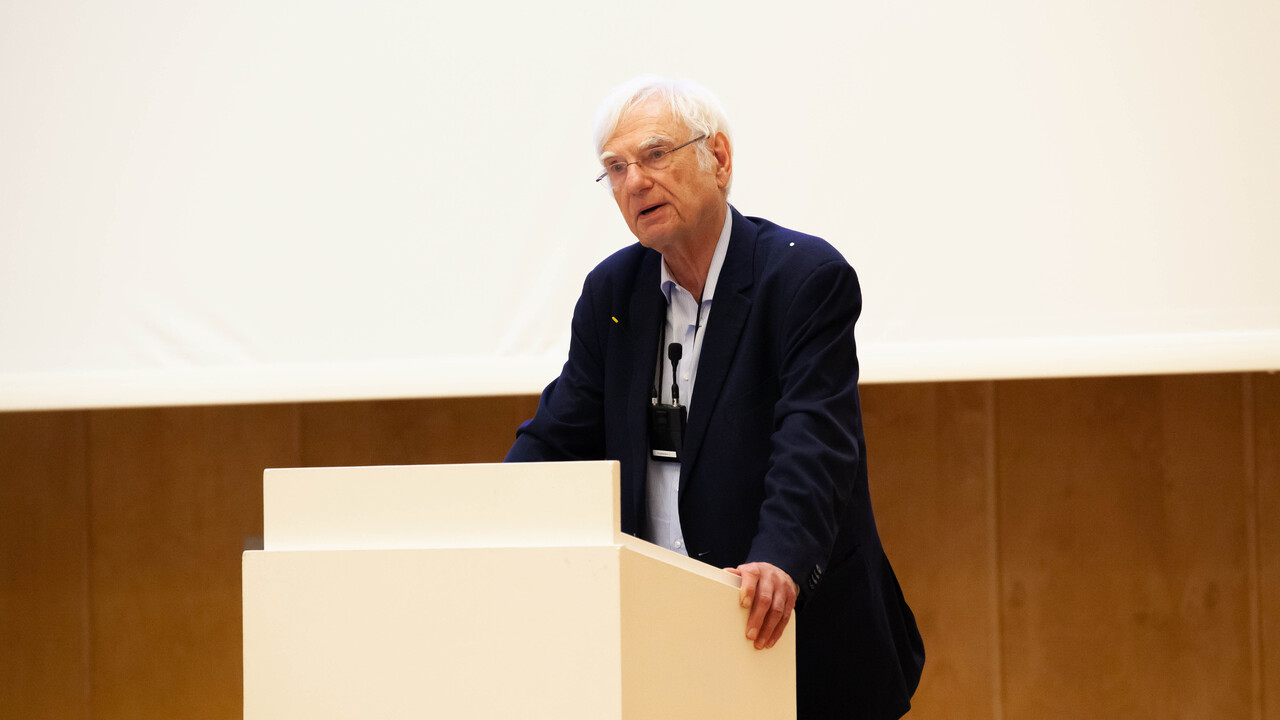 Prof. Dr. Dr. h. c. Dieter Birnbacher steht vor dunkler Holzvertäfelung und heller Projektionsfläche hinter einem hellen Pult, auf das er seine Hände stützt. Er ist bekleidet mit einem dunkelblauen Anzug und einem hellblaues Hemd. Ein trägt eine Brille und ein umgehängtes Funkmikrofon.