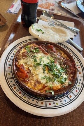 Das israelische Gericht Shakshuka: Überbackenes Eiergericht in einer Tonschale mit Kräutern und Fladenbrot auf einem Tablett, daneben eine Flasche Cola.
