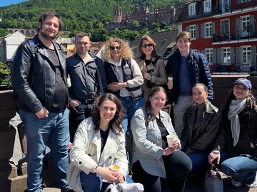 Eine Gruppe von neun Personen auf der Alten Brücke in Heidelberg, im Hintergrund rechts ein rotes Haus und die Ruine des Heidelberger Schlosses.