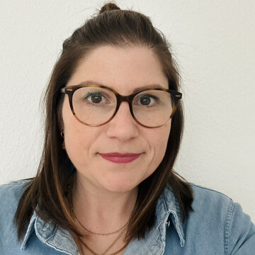 Frau mit braunen, schulterlangen Haaren und Brille, trägt eine hellblaue Jeansbluse und zwei goldene Ketten