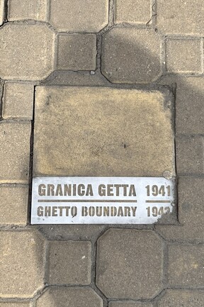 Pflastersteine auf einem Gehsteig. In der Mitte ein größerer quadratischer Stein mit der Aufschrift "GRANICA GETTA 1941 – GHETTO BOUNDARY 1942"