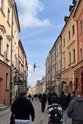 Eine Straße in der Altstadt Lublin. Zu beiden Seiten stehen bunte Häuser, dazwischen laufen einige Menschen. Zwischen den Häuserfronten hängt die Figur eines Seiltänzers in der Luft. 
