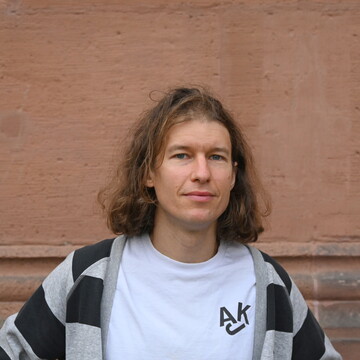 Richard Rohrmoser steht vor einer rötlich-braunen Wand. Er trägt ein weißes Shirt, darüber eine grau-Schwarz gestreifte Stoffjacke. Er hat schulterlanges braun-blondes Haar. 