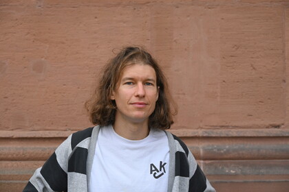 Richard Rohrmoser steht vor einer rötlich-braunen Wand. Er trägt ein weißes Shirt, darüber eine grau-Schwarz gestreifte Stoffjacke. Er hat schulterlanges braun-blondes Haar. 