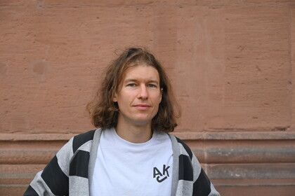 Richard Rohrmoser steht vor einer rötlich-braunen Wand. Er trägt ein weißes Shirt, darüber eine grau-Schwarz gestreifte Stoffjacke. Er hat schulterlanges braun-blondes Haar. 