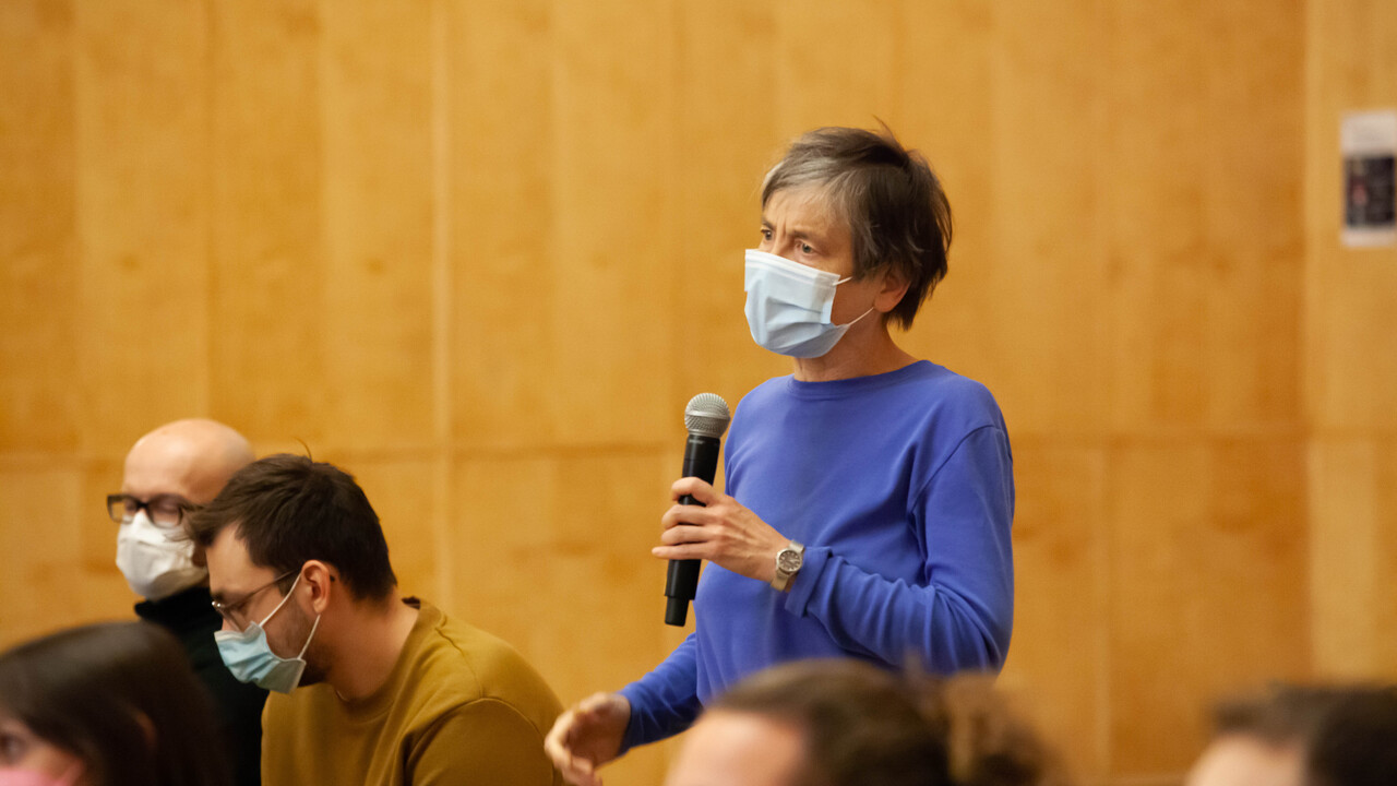 Frage aus dem Publikum: Eine stehende Person mit Corona-Atmschutzmaske hält ein Mikrofon in der linken Hand mit etwas Abstand auf Kinnhöhe und blickt nach links aus dem Bild. Um die Person herum befinden sich 5 weitere Menschen auf Stühlen sitzende Menschen mit Atemschutzmasken, teilweise außerhalb der Bildschärfe angeschnitten im Kameraausschnitt. Den Hintergrund bildet eine Holzvertäfelung.