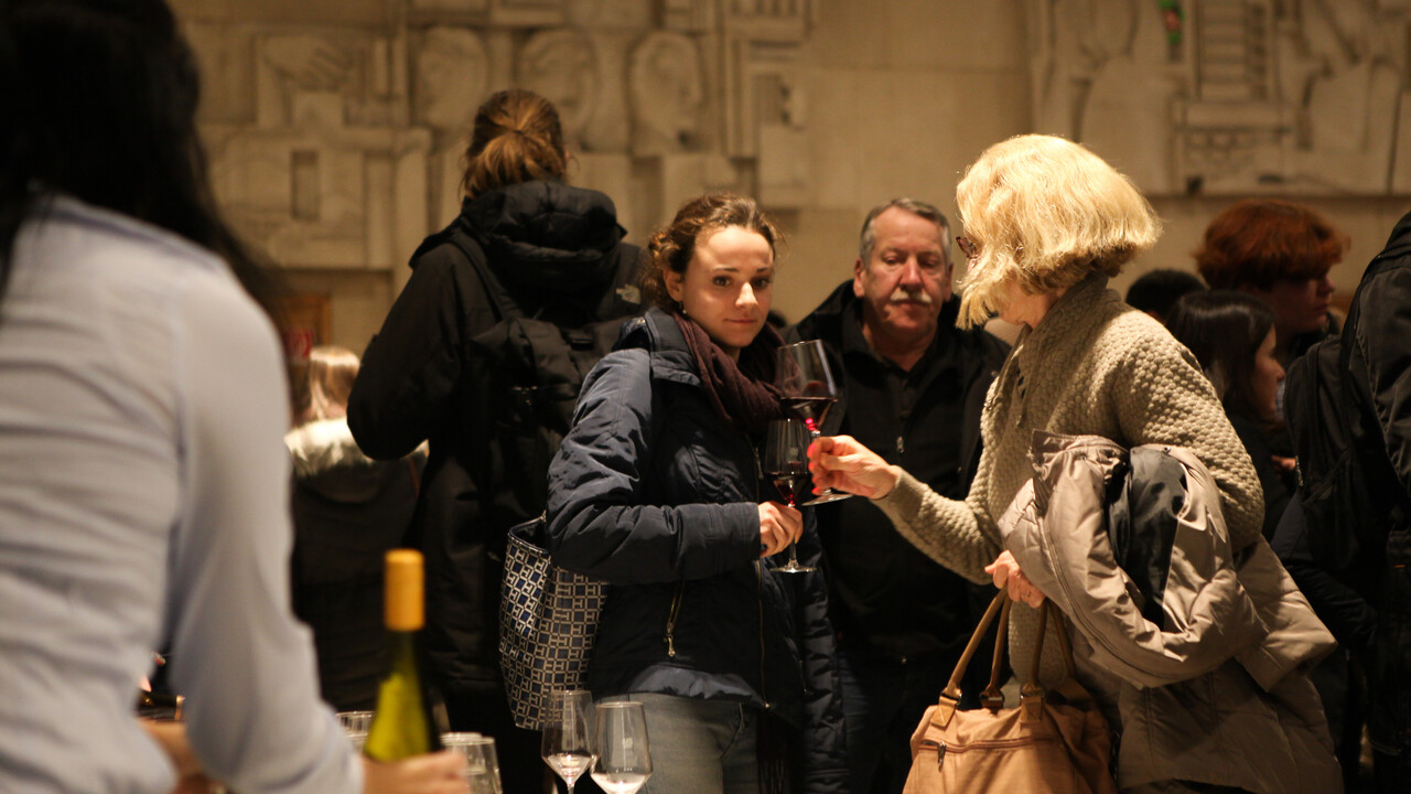 Im Vordergrund von hinten schlanke Weingläser und eine Flasche, die gerade bereitgestellt werden. Im Hintergrund Menschen winterlich gekleidete Menschen im Gespräch mit Gläsern in den Händen vor einer Wand mit künstlerisch gestalteter heller Wand. Eine jüngere und ältere Damen sind gerade im Begriff anzustoßen.