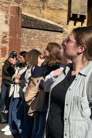 7 nach links blickende Frauen vor einem historischen Gemäuer.