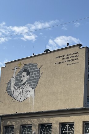 Ein Mural auf einer Hauswand in Warschau: Ein Mann hält mit erhobenem Arm eine Narzisse. Daneben ein Zitat von Marek Edelmann: "NIENAWIŚĆ JEST LATWA. MILOŚĆ WYMAGA WYSIŁKU I POŚWIĘCENIA." ("Hass ist leicht. Liebe erfordert Anstrengung und Opfer.") 