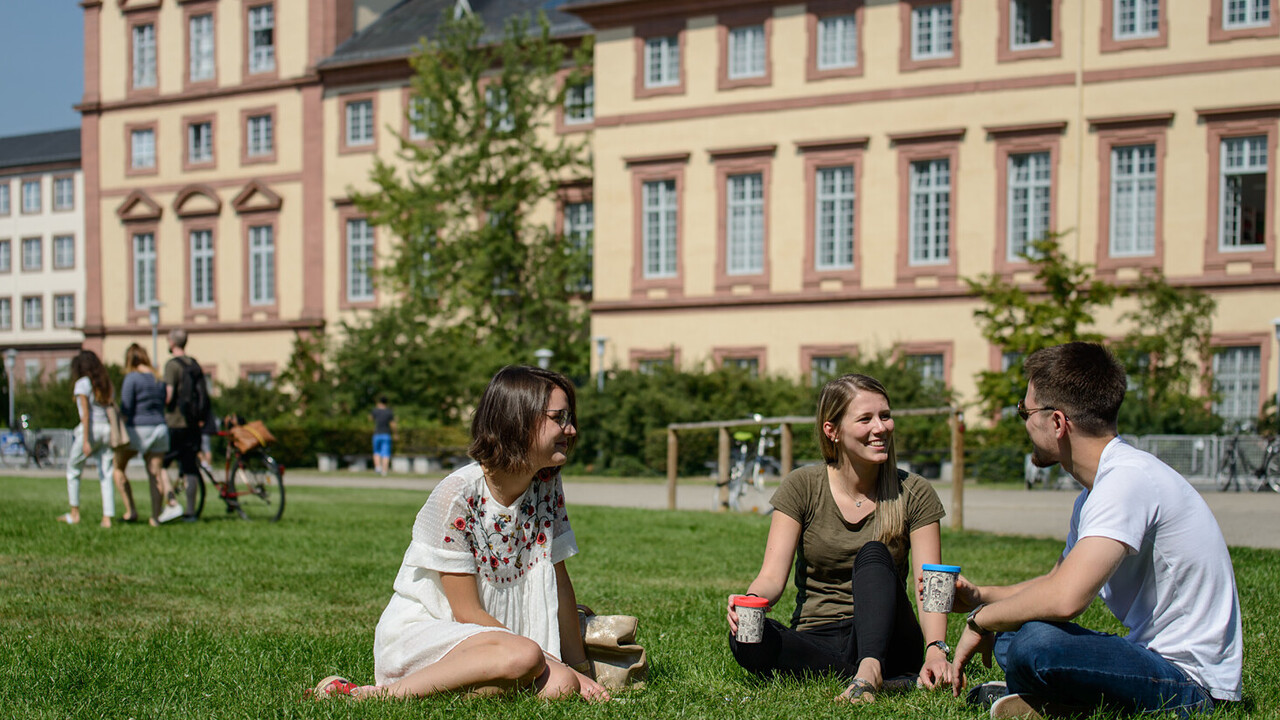 Zwei Studentinnen und ein Student sitzen auf einer grünen Wiese. Sie tragen frühlingshafte Kleidung und halten Kaffee-To-Go-Becher in ihren Händen.