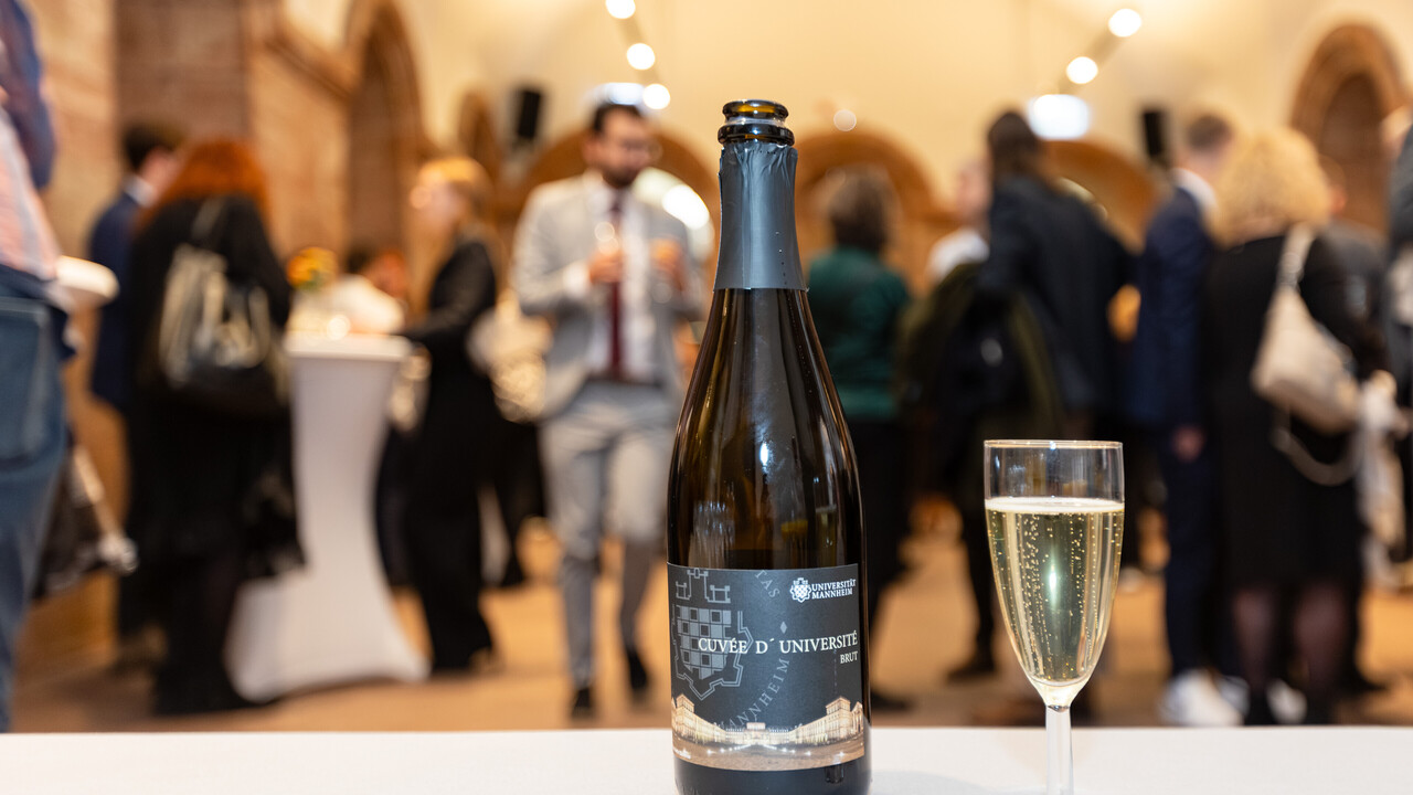 Im Vordergrund steht eine Flasche Sekt mit dem Loo der Uni Mannheim, daneben ein gefülltes Sektglas. Im Hintergrund sind verschwommen Gäste zu erkennen.