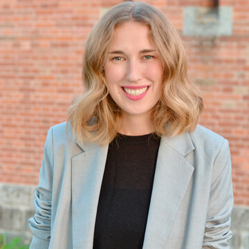 Vivian Seidel trägt ein schwarzes Oberteil und darüber einen hellen blau-grünen Blazer. Sie hat etwa schulterlanges blondes Haar und lächelt. 