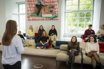 Eine Frau steht mit dem Rücken zur Kamera und spricht zu einer Gruppe von Studierenden, die in einem hellen, modernen Seminarraum auf gestuften Sitzflächen und Sesseln sitzen. Einige halten Tablets oder Notizblöcke in der Hand und hören aufmerksam zu.