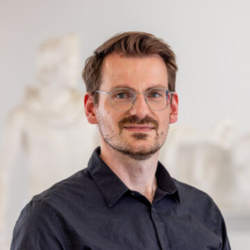 Jonas Brosig trägt ein dunkles Hemd, eine silberne Brille und einen Bart. 