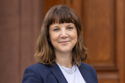 Frau mit schulterlangem braunem Haar trägt weißes T-Shirt und dunkelblauen Blazer vor Holzwand