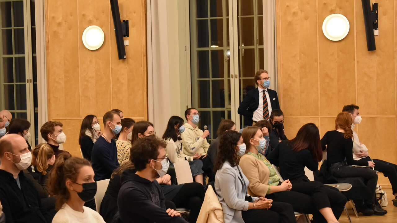 Blick von schräg vorne auf das herbstlich gekleidete Publikum, das mit Corona-Atemschutzmasken auf leicht auseinandergezogenen Stuhlreihen sitzt, von denen die meisten besetzt sind. Im Hintergrund die durch Fenster durchbrochene holzvertäfelte Außenwand, an der rechteckige Lautsprecher und runde Leuchten angebracht sind. An einem der Fenster lehnt ein Mann mit weißem Hemd und dunklen Anzug nebst Krawatte mit Brille, der ihm am nächsten sitzende Zuschauer hät ein Mikrofon in der Hand.