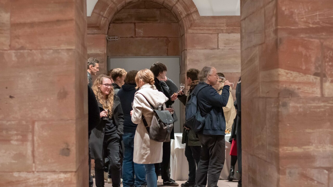 Blick durch einen Sandsteinsbogen beim Empfang in den „Katakomben“ auf herbstlich gekleidete Personen, die sich mit Gläsern in den Händen unterhalten oder daraus trinken.