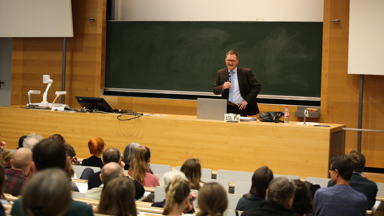 Blick aus dem Publikum auf Prof. Dr. Philipp Gassert bei seinem Grußwort als Dekans der Philosophischen Fakultät. Man sieht die zur dunkelgrünen Tafel und Pult abfallenden Sitzplatzreihen mit den Köpfen des Auditoriums von hinten. Prof. Gassert steht in hellem Hemd mit hellblauer, von schmalen weißen und dunklen schrägen Streifen gekreuzter Krawatte und braunem Sakko mit rötlichem sich kreuzendem Strichmuster gekleidet zwischen Pult und Tafel. Die Tafel ist eingebettet in eine Holzvertäfelung, rechts, links und oberhalb sich weiße Flächen im Anschnitt zu sehen.