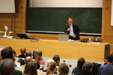 Blick aus dem Publikum auf Prof. Dr. Philipp Gassert bei seinem Grußwort als Dekans der Philosophischen Fakultät. Man sieht die zur dunkelgrünen Tafel und Pult abfallenden Sitzplatzreihen mit den Köpfen des Auditoriums von hinten. Prof. Gassert steht in hellem Hemd mit hellblauer, von schmalen weißen und dunklen schrägen Streifen gekreuzter Krawatte und braunem Sakko mit rötlichem sich kreuzendem Strichmuster gekleidet zwischen Pult und Tafel. Die Tafel ist eingebettet in eine Holzvertäfelung, rechts, links und oberhalb sich weiße Flächen im Anschnitt zu sehen.