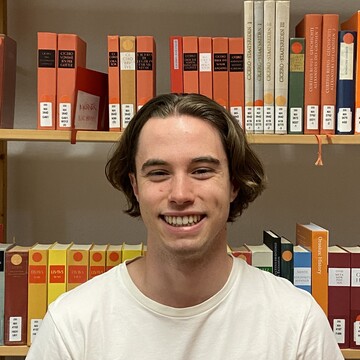 Philipp Hagnerhat mittellange braune Haare. Er trägt ein weißes T-Shirt und steht vor einem Bücherregal.
