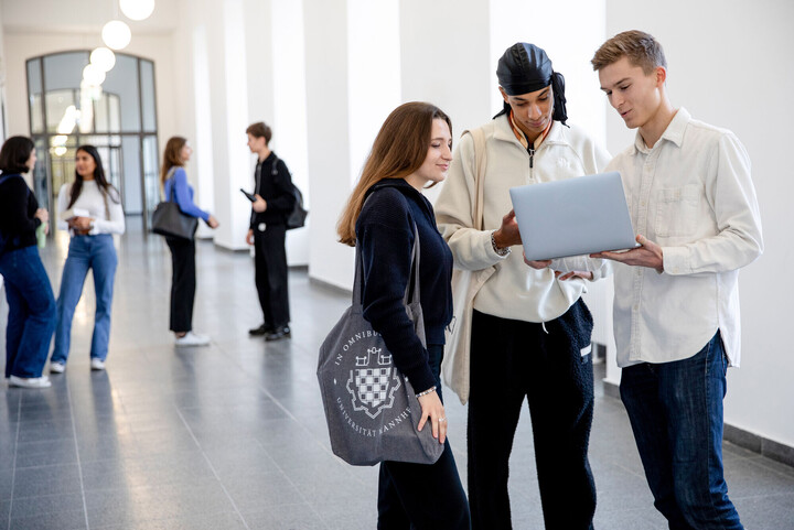 Eine Gruppe von Studierenden steht im Flur der Uni und unterhalten sich. Im Vordergund betrachten eine Studentin und zwei Studenten etwas in einem Laptop.