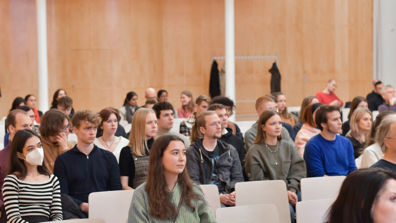 Blick von schräg vorne auf das herbstlich gekleidete Publikum. Einige Plätze sind frei, eine Frau links im Bild am Ende einer vollbesetzten Stuhlreihe trägt eine Atemschutzmaske. Im Hintergrund ist eine holzvertäfelte Wand mit einem Garderobenständer hinter einer hellen Säule, an dem 3 Jacken oder Mäntel hängen.