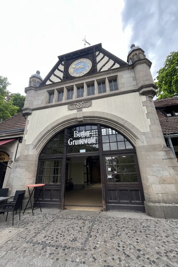 Historisches Eingangsgebäude des Bahnhofs Berlin-Grunewald mit großer Rundbogenfassade und markanter Uhr unter dem Giebel. Vor dem Gebäude liegt ein gepflasterter Vorplatz, links ein kleines Café.