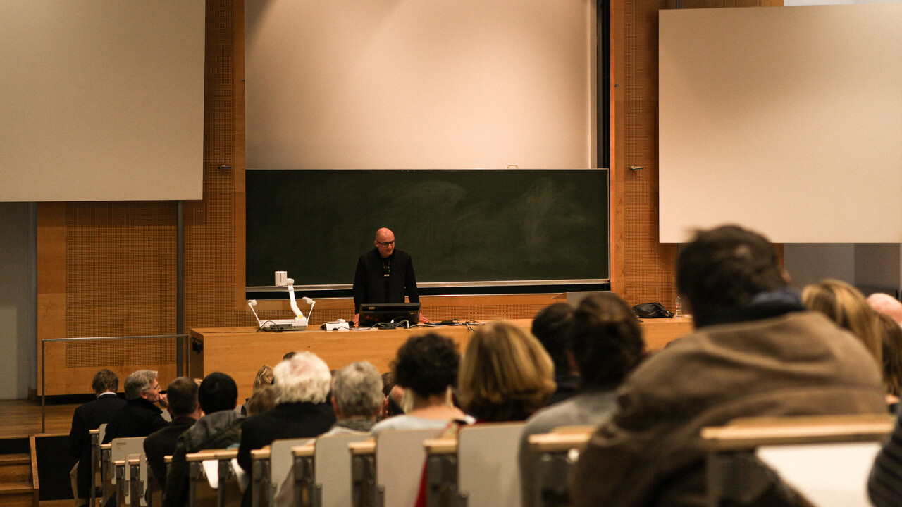 Blick aus dem Publikum auf Prof. Dr. Wolfgang Freitag bei der Begrüßung der Gäste. Man sieht die zur dunkelgrünen Tafel und Pult abfallenden Sitzplatzreihen mit den Köpfen des Auditoriums von hinten. Prof. Freitag steht in schwarz gekleidet zwischen Pult und Tafel. Die Tafel ist eingebettet in eine Holzvertäfelung, rechts, links und oberhalb befinden sich weiße Projektionsflächen.