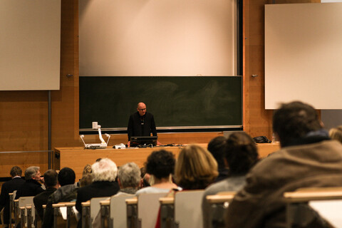 Blick aus dem Publikum auf Prof. Dr. Wolfgang Freitag bei der Begrüßung der Gäste. Man sieht die zur dunkelgrünen Tafel und Pult abfallenden Sitzplatzreihen mit den Köpfen des Auditoriums von hinten. Prof. Freitag steht in schwarz gekleidet zwischen Pult und Tafel. Die Tafel ist eingebettet in eine Holzvertäfelung, rechts, links und oberhalb befinden sich weiße Projektionsflächen.