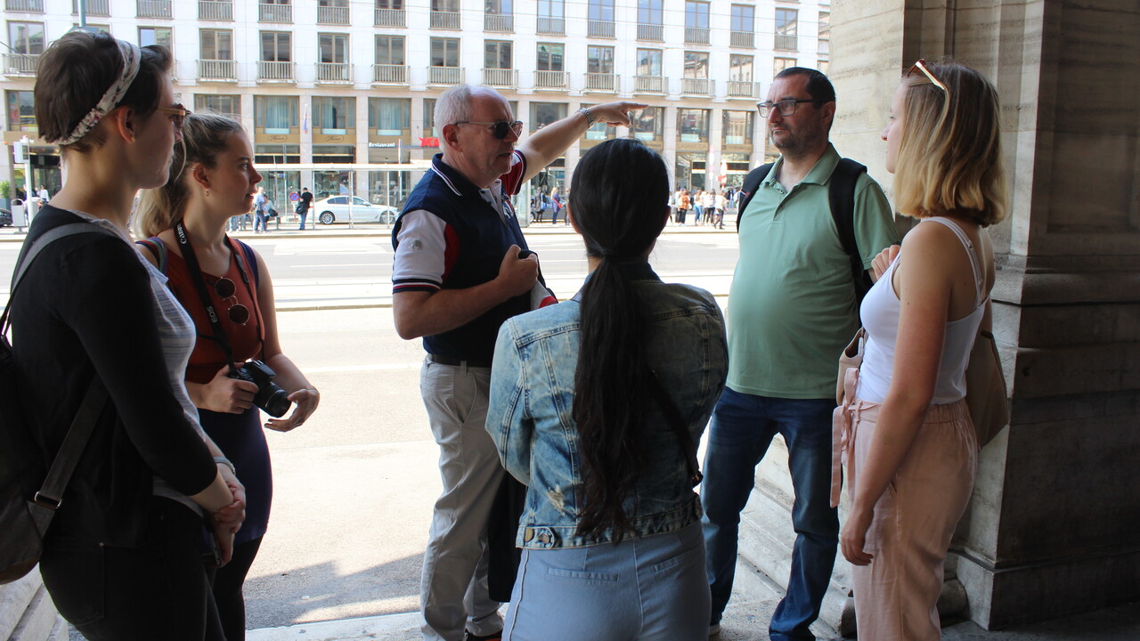6 Personen in Freizeitkleidung neben einer Säule oder in der vorgelagerten Arkade eines Gebäudes im Schatten stehend. Ein zentral zwischen allen anderen stehender älterer Herr deutet mit einem Arm nach rechts auf einen Punkt außerhalb des Bildes. Im Hintergrund sind sonnenbeschienen eine Straße mit Haltestelle und dahinter die weiße Fassade eines großen Gebäudes mit vielen Fenstern zu sehen.