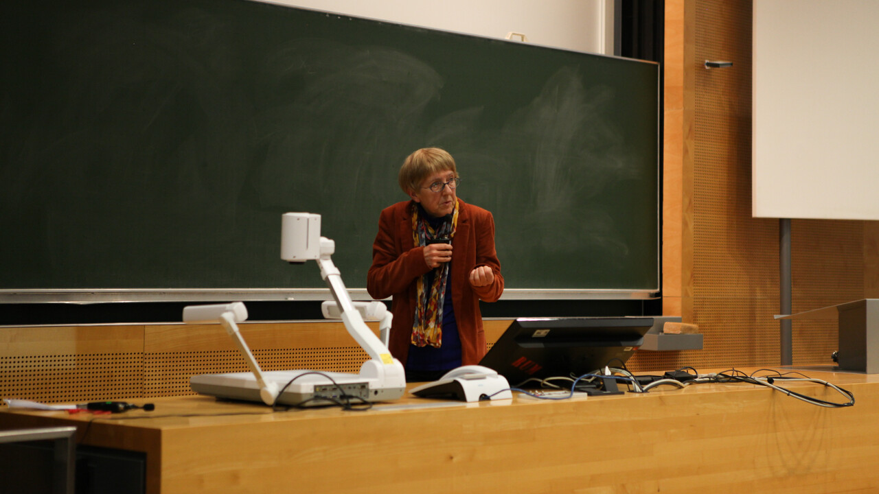 Prof. Dr. Ursula Wolf steht vor einer grünen Tafel hinter einem Pult mit Monitor und anderen technischen Accessoires. Sie hält mit einer Hand ihr umgehängtes Funktmikro etwas mehr in Mundnähe und gestikuliert mit der anderen Hand eine greifende Bewegung. Prof. Wolf trägt eine Brille und ist mit einem rötlichen Jaket und einem farbenfrohen Schal gekleidet.