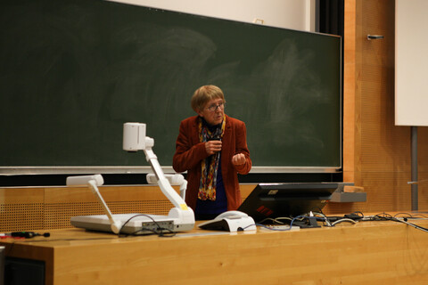 Prof. Dr. Ursula Wolf steht vor einer grünen Tafel hinter einem Pult mit Monitor und anderen technischen Accessoires. Sie hält mit einer Hand ihr umgehängtes Funktmikro etwas mehr in Mundnähe und gestikuliert mit der anderen Hand eine greifende Bewegung. Prof. Wolf trägt eine Brille und ist mit einem rötlichen Jaket und einem farbenfrohen Schal gekleidet.