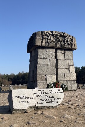 Die Gedenkstätte des ehemaligen Vernichtungslagers Treblinka: In der Mitte befindet sich ein meterhoher Stein. Davor steht eine kleinere Steintafel mit der Aufschrift "Nie wieder" in verschiedenen Sprachen. Im Hintergrund sieht man Grabsteine und freie Fläche umgrenzt von Wald. 