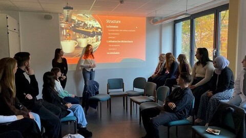 Eine Gruppe Studierender steht vor ihrer Präsentation und erklärt den ausgearbeiteten Lehrstoff. Schüler*innen und weitere Studierende sitzen im Publikum und folgen aufmerksam der Vorstellung.