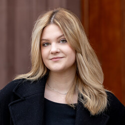 Porträt von Emilia Toth. Emilia Toth trägt einen schwarzen Mantel und ein schwarzes Oberteil. Sie hat blonde Haare, die sie offen trägt und die ihr über die Schultern reichen. Um den Hals trägt sie eine dezente silberne Kette. Sie schaut offen in die Kamera.