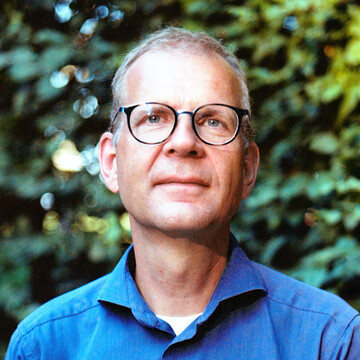 Dr. Christoph Böhm. Er hat eine Brille auf und trägt ein dunkelblaues Hemd. Den Hintergrund bilden grüne Blätter von Pflanzenbewuchs.